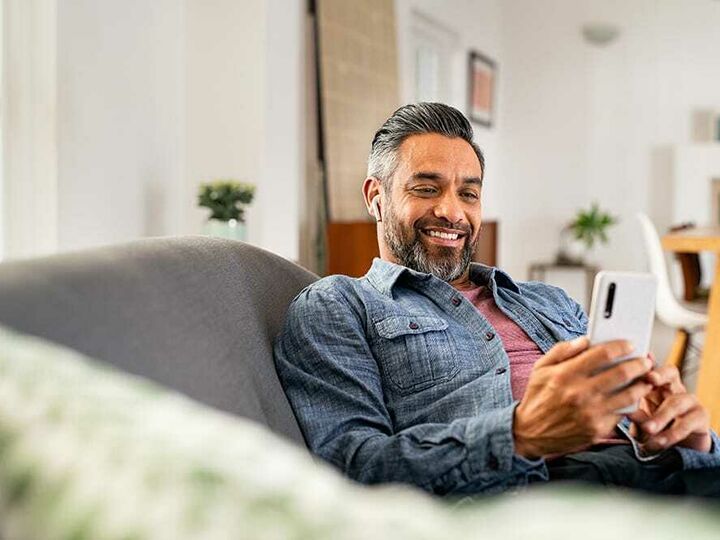 Smiling Exchange Bank customer relaxing at home on the couch, reviewing bank account online on cell phone through Exchange Bank mobile app.
