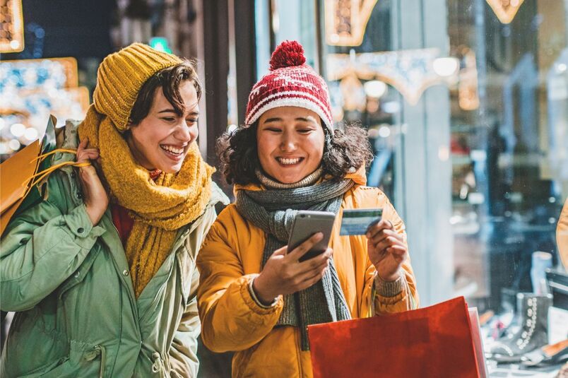 Two women wearing winter coats smile while shopping, one holding a smartphone and a bank card, representing secure everyday banking.