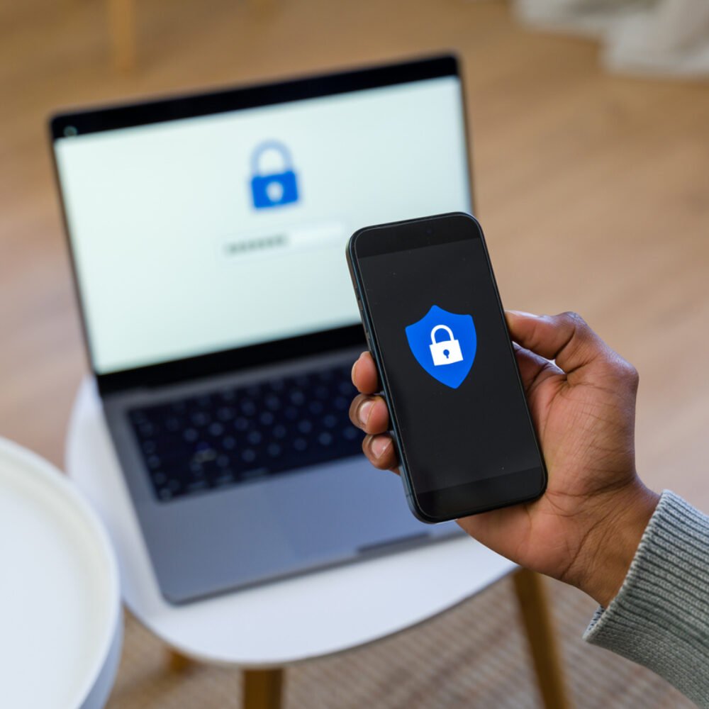 Person holding a phone with an image of secure lock with a laptop displaying the same image, slightly out of focus in the background