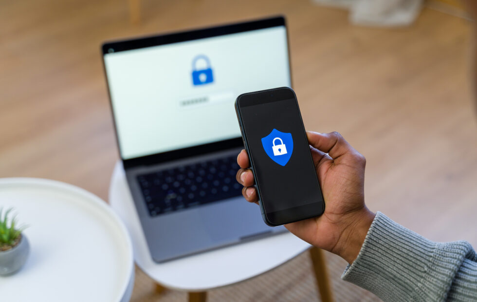 Person holding a phone with an image of secure lock with a laptop displaying the same image, slightly out of focus in the background